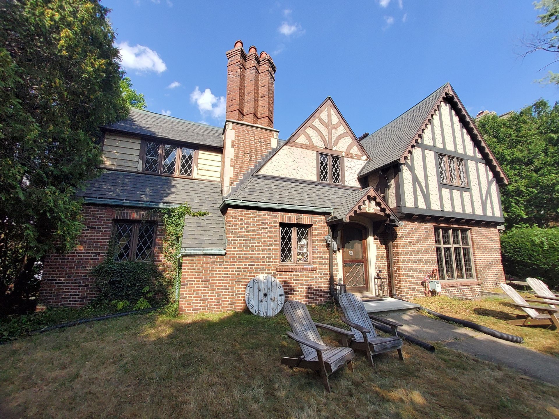 Tudor-style brick house with a dark roof and decorative trim. Two Adirondack chairs sit on the lawn.