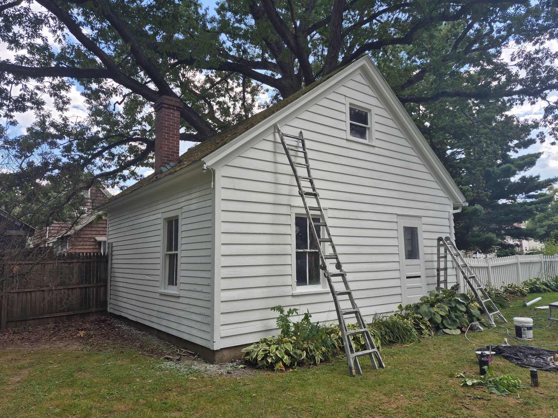 White house with ladders, a brick chimney, and a picket fence. Overgrown with foliage, cloudy sky.
