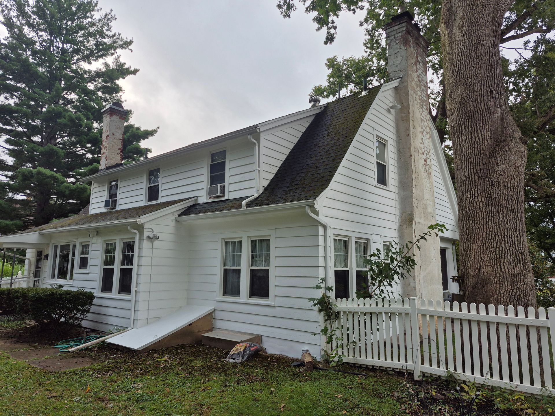White two-story house with black roof, chimney, and white picket fence. A large tree is on the right side.