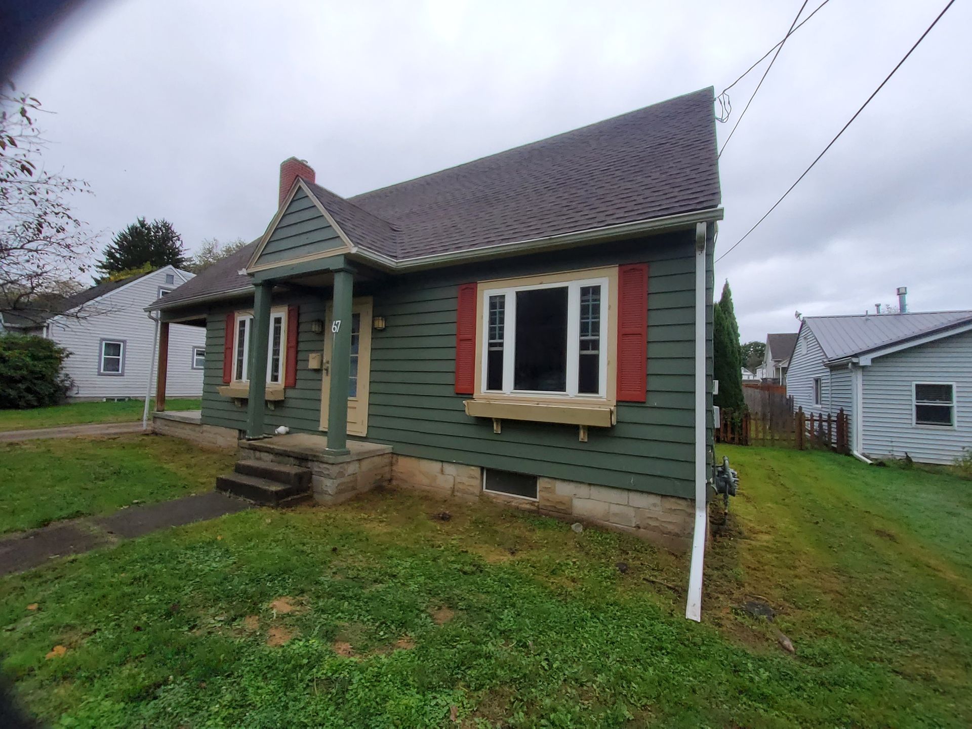 Green house with red shutters, bay window, and dark roof on a grassy lot under a cloudy sky.