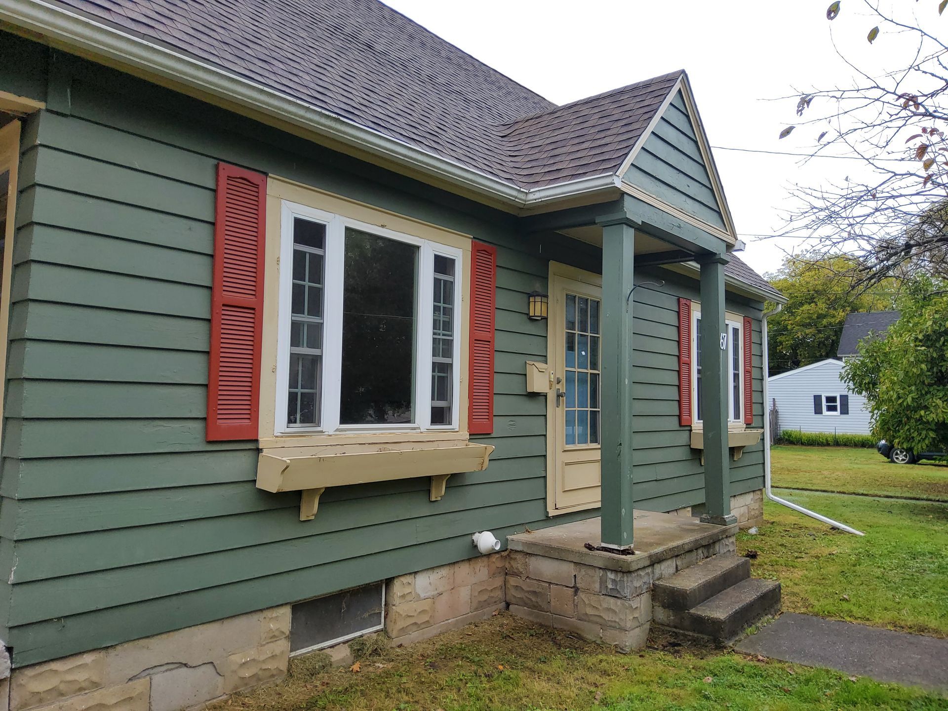 Green house with red shutters, beige trim, steps, and a porch.
