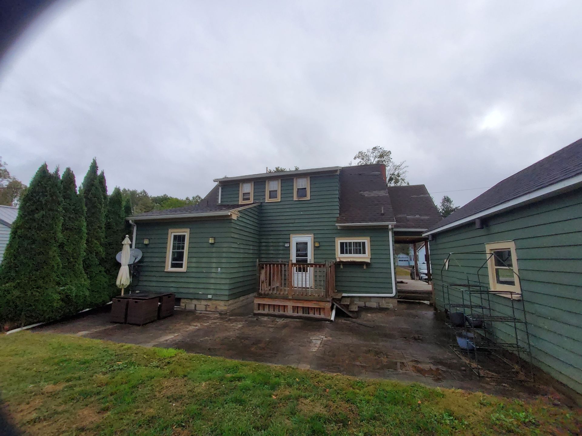 Back of a green two-story house with a damaged roof and a small wooden deck, under an overcast sky.