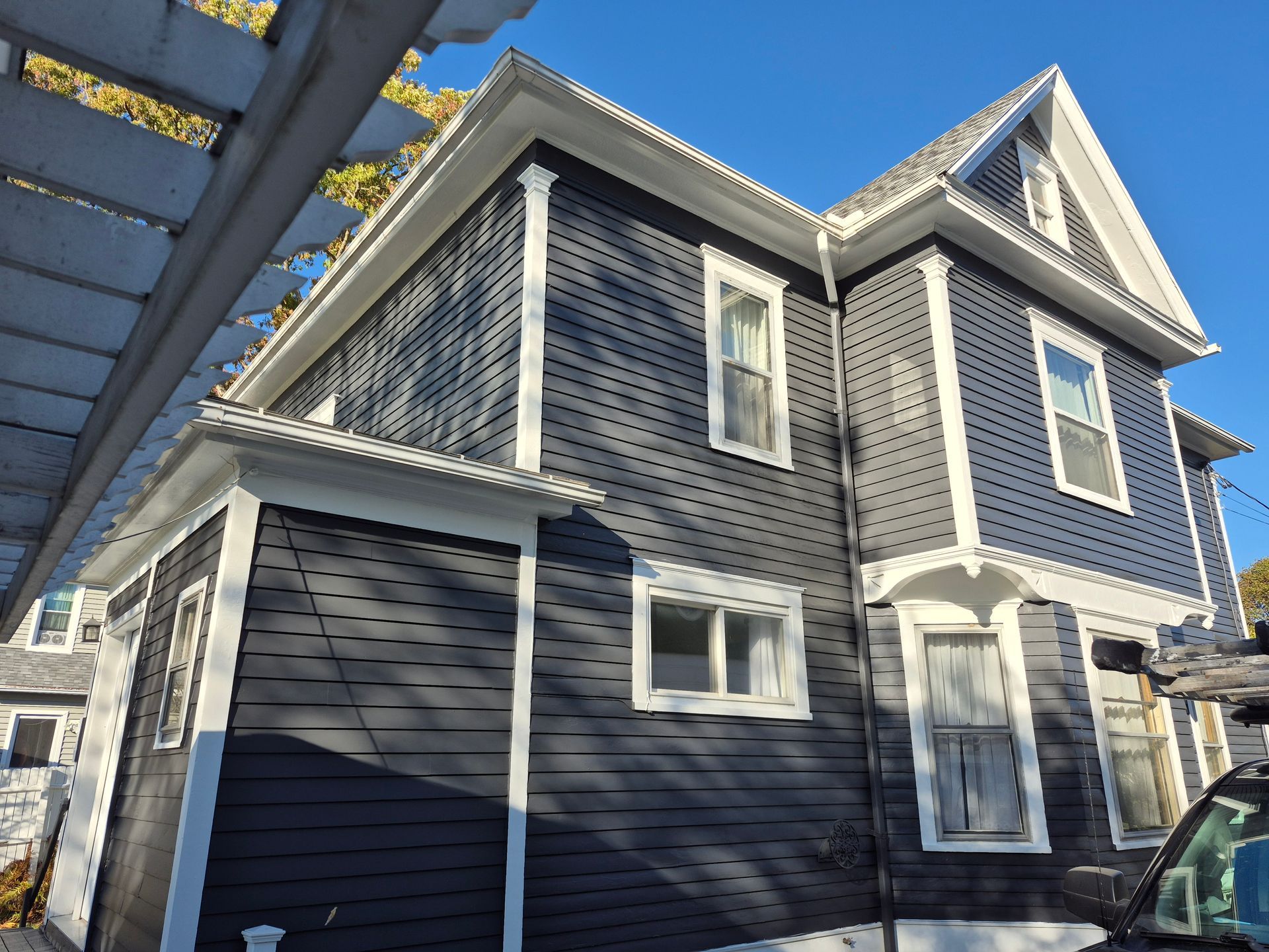 Two-story house with dark gray siding, white trim, and a pergola against a blue sky.