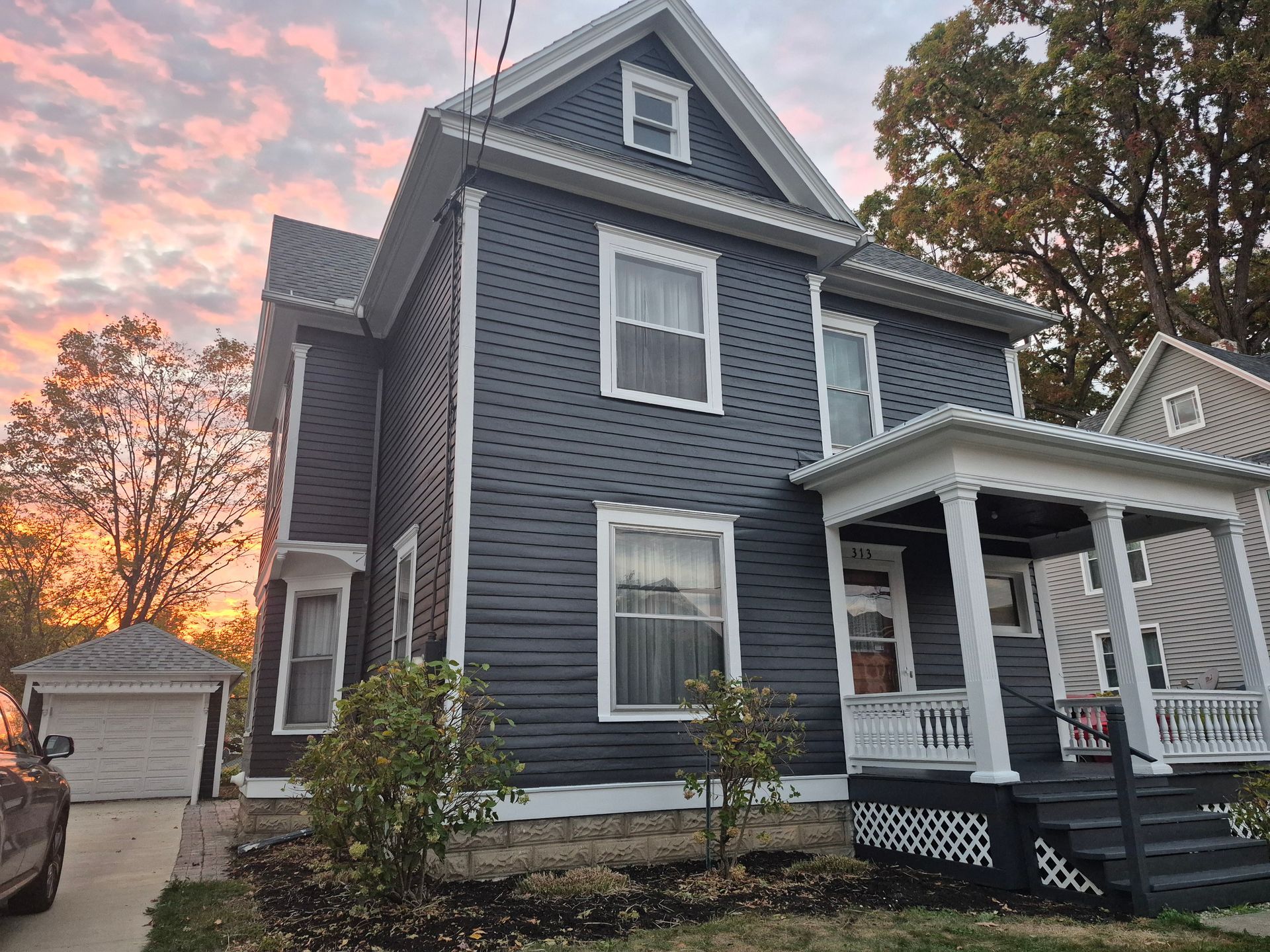 Two-story house, dark gray siding with white trim, porch, and a garage in the background. Sunset sky.