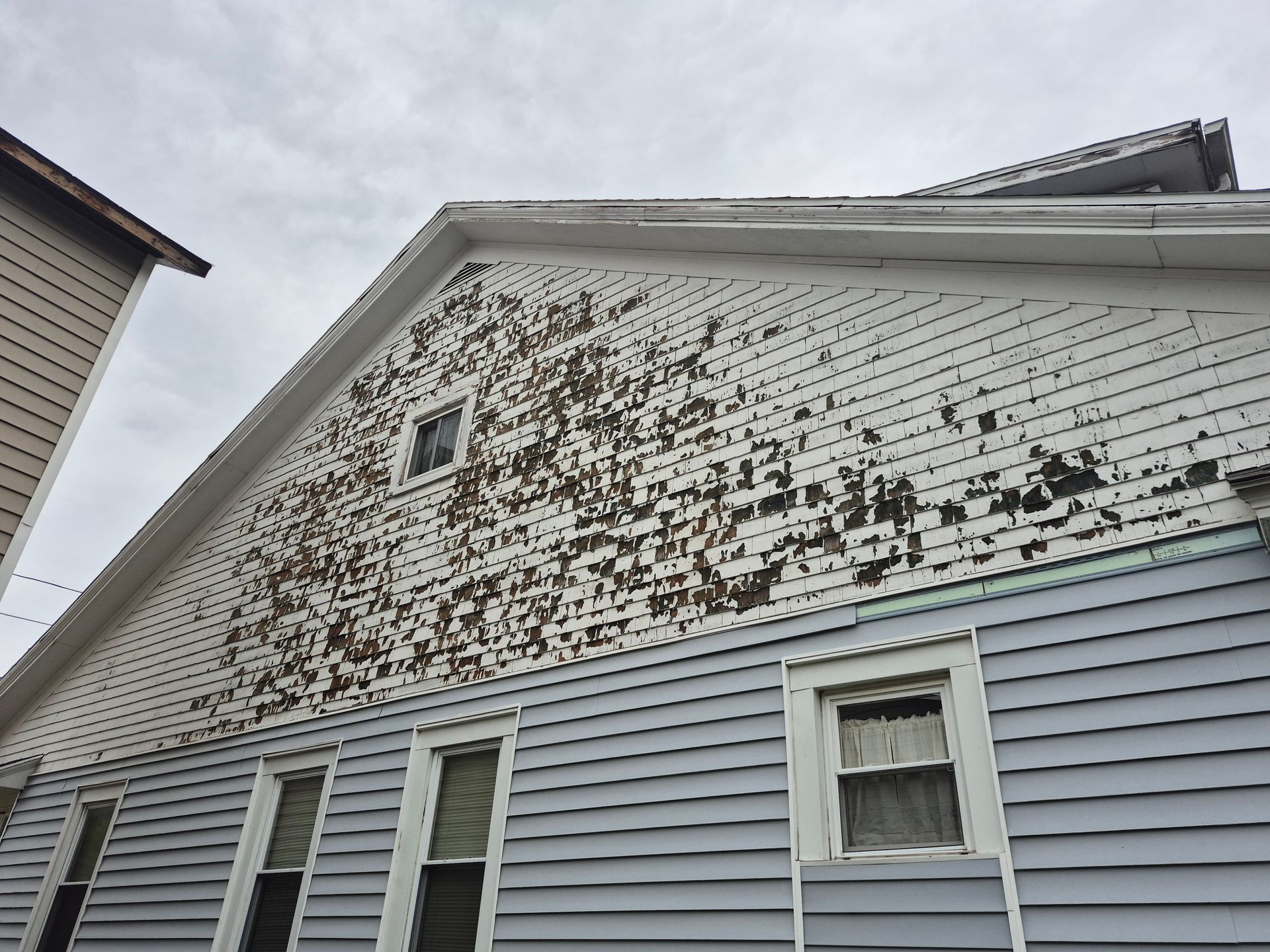 House exterior: peeling paint on siding and gable, blue siding below, windows. Overcast sky.