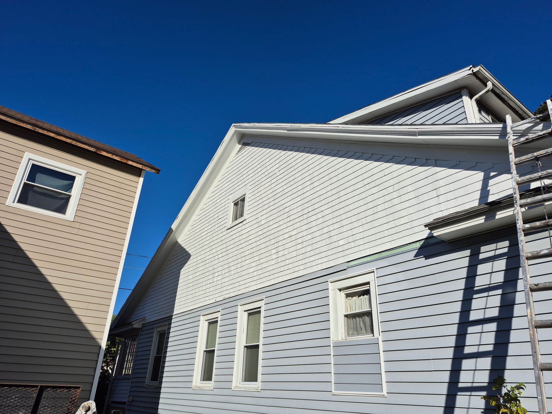 Side of a two-story blue and white house with a ladder propped up. A beige building is to the left. Bright blue sky.