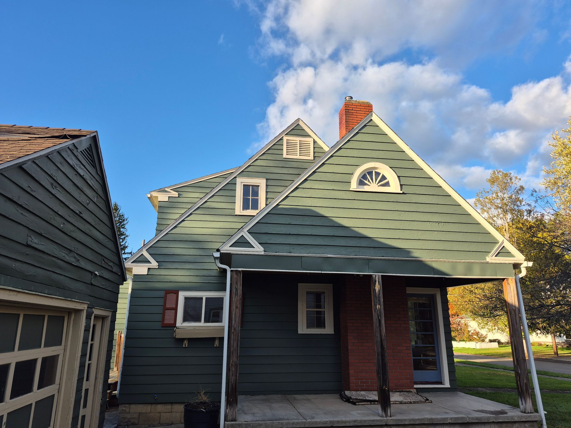 Green house with white trim, brown porch, and a brick chimney under a blue sky with clouds.