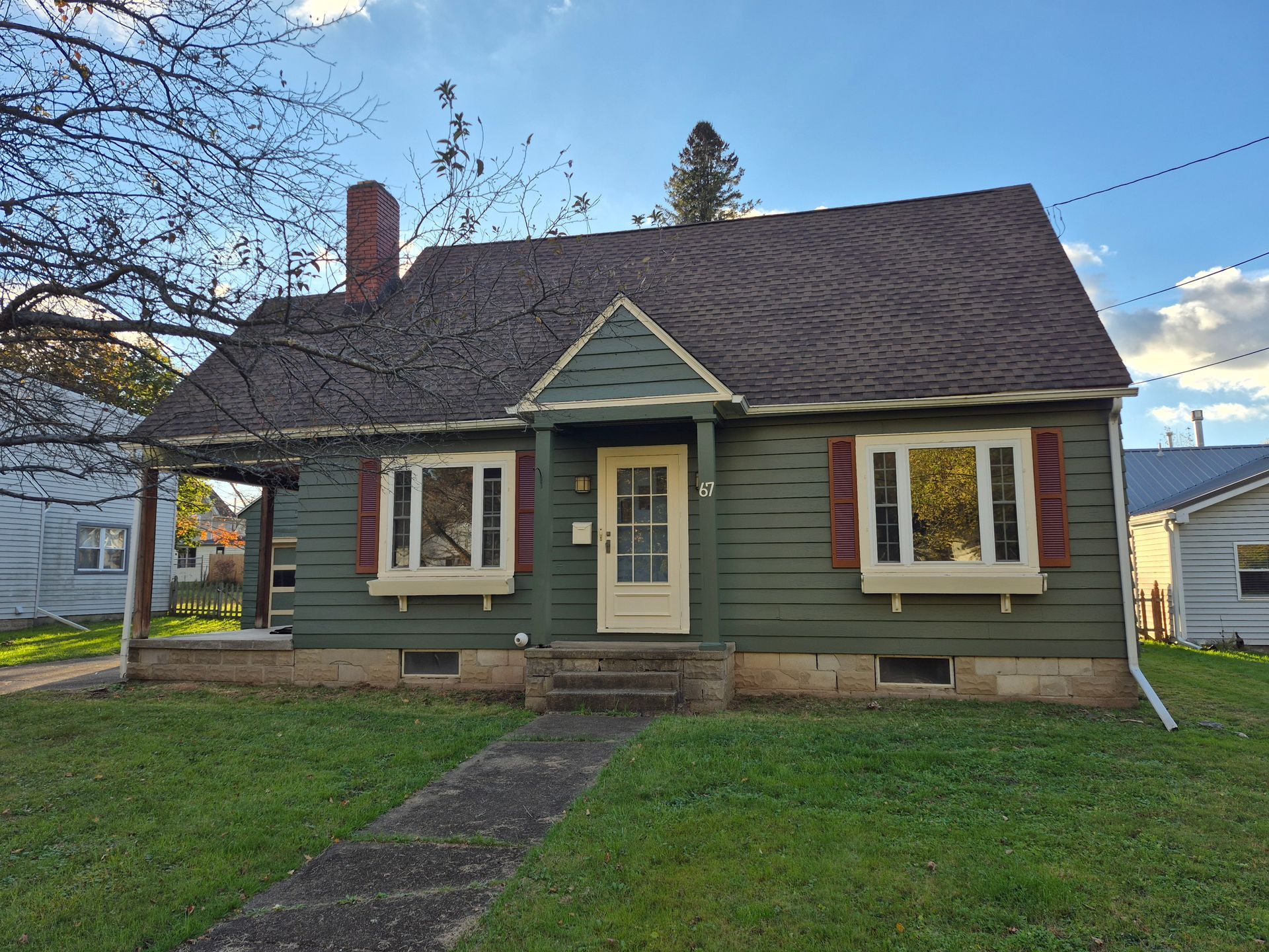 Green bungalow with red shutters, brown roof, and brick chimney on a grassy lawn.