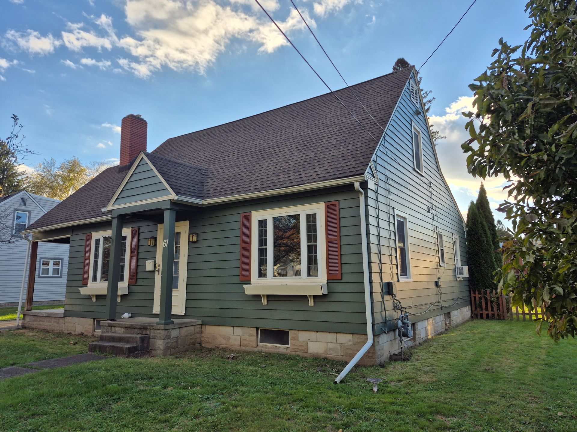 Green house with red shutters, chimney, and a small porch on a grassy lawn under a cloudy sky.