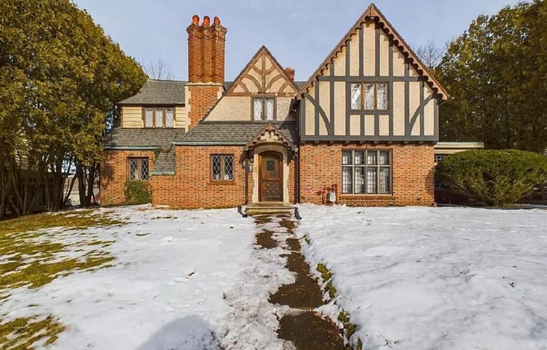 Tudor-style brick house with snow-covered yard and walkway. Brown and white accents, arched doorway, and chimney.