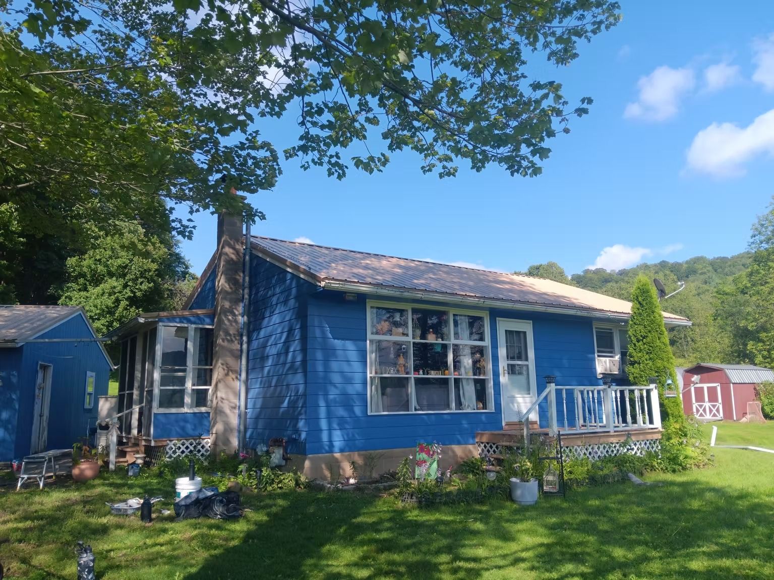 Blue house with white-framed windows, porch, and chimney, under a blue sky, surrounded by trees and grass.