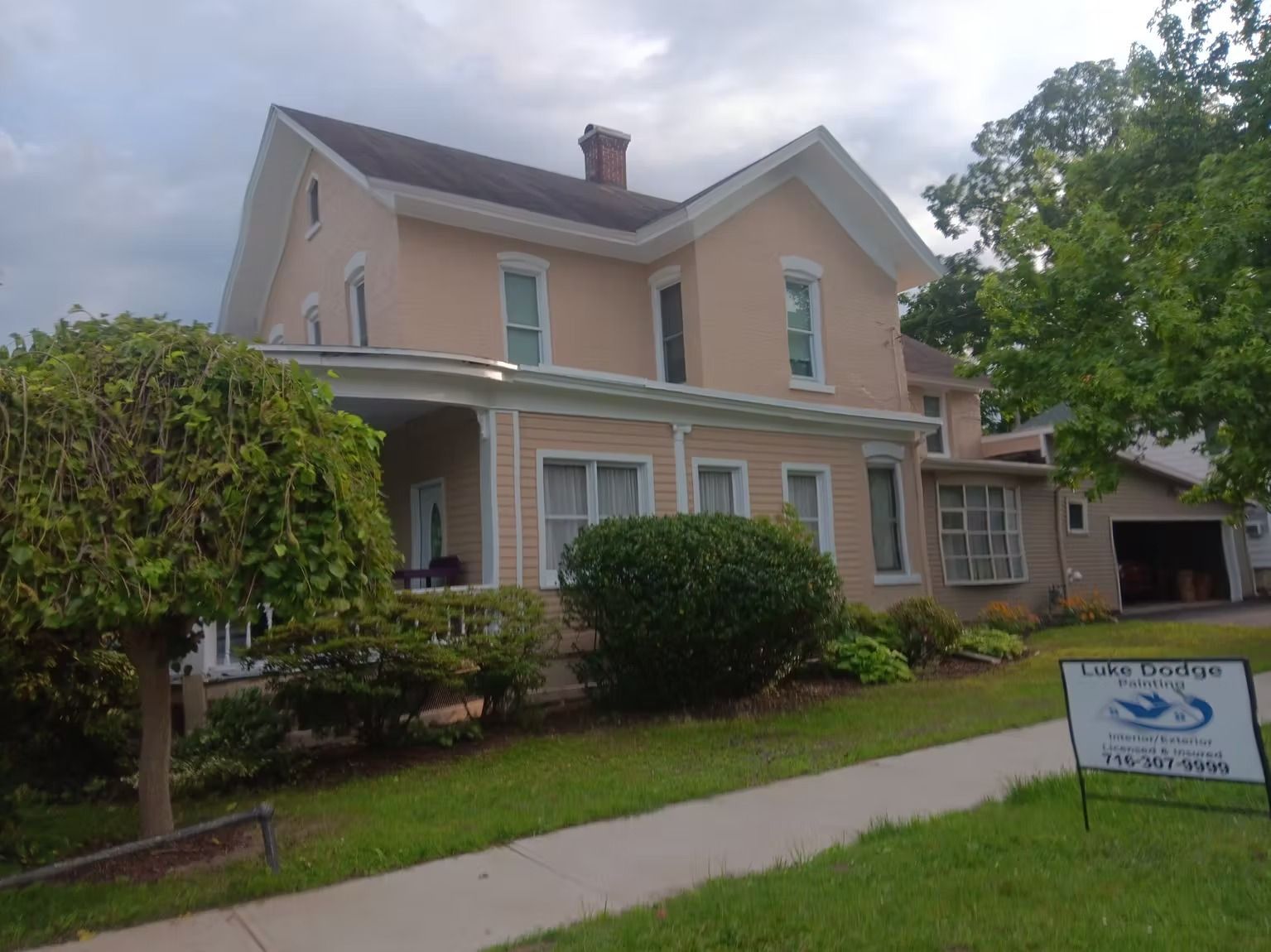 Two-story house with tan siding, porch, and a green lawn. A sign for 