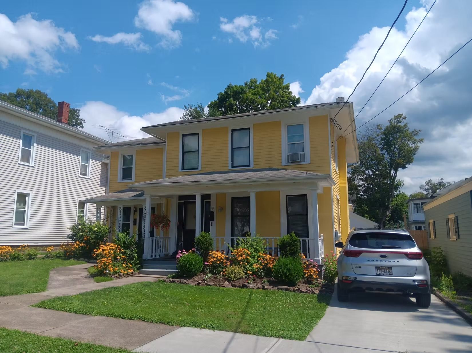 Yellow two-story house with porch, white trim, and a car parked in driveway under a blue sky.