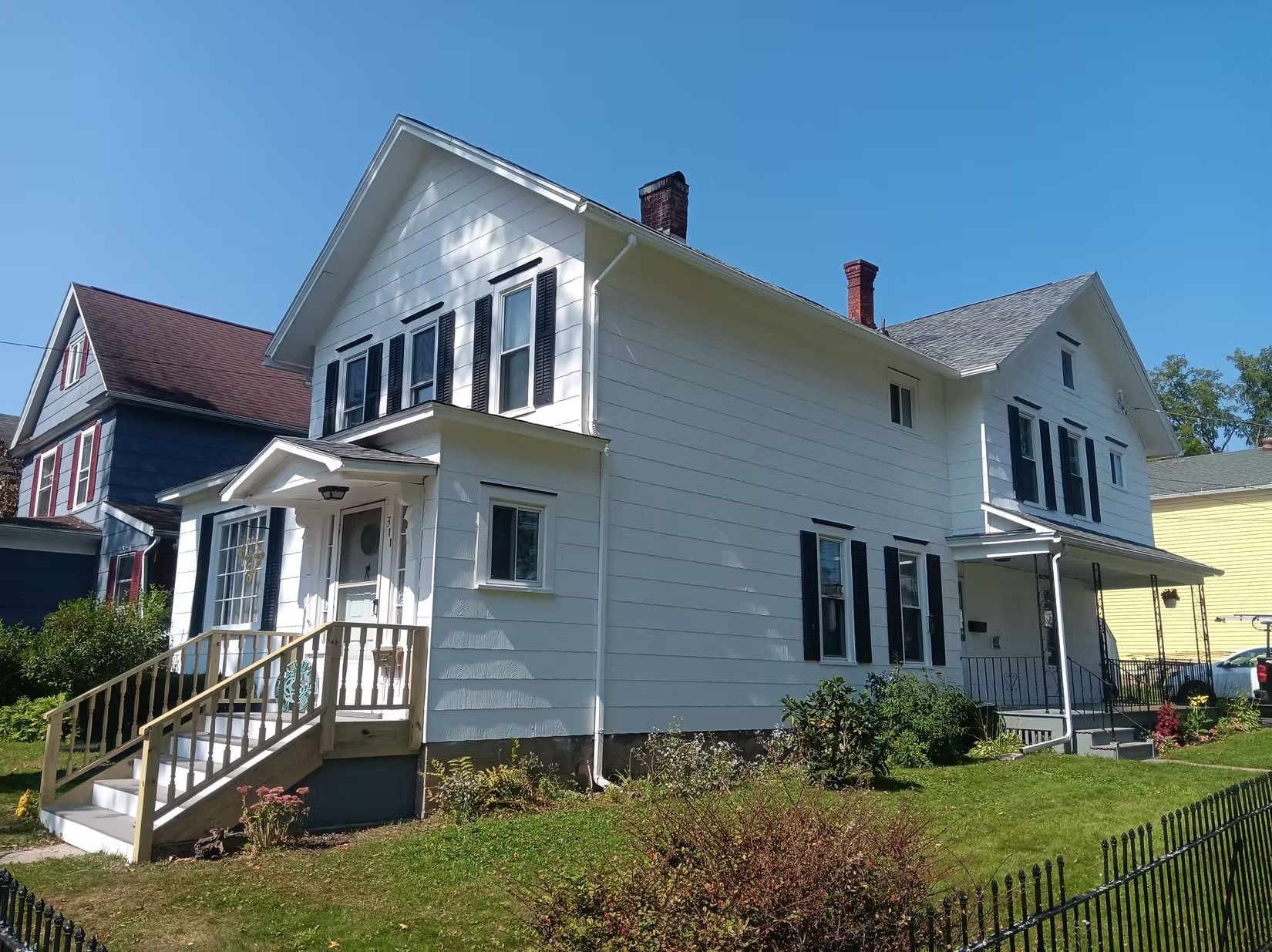 White two-story house with black shutters, wooden porch, and a brick chimney. Green grass and blue sky.