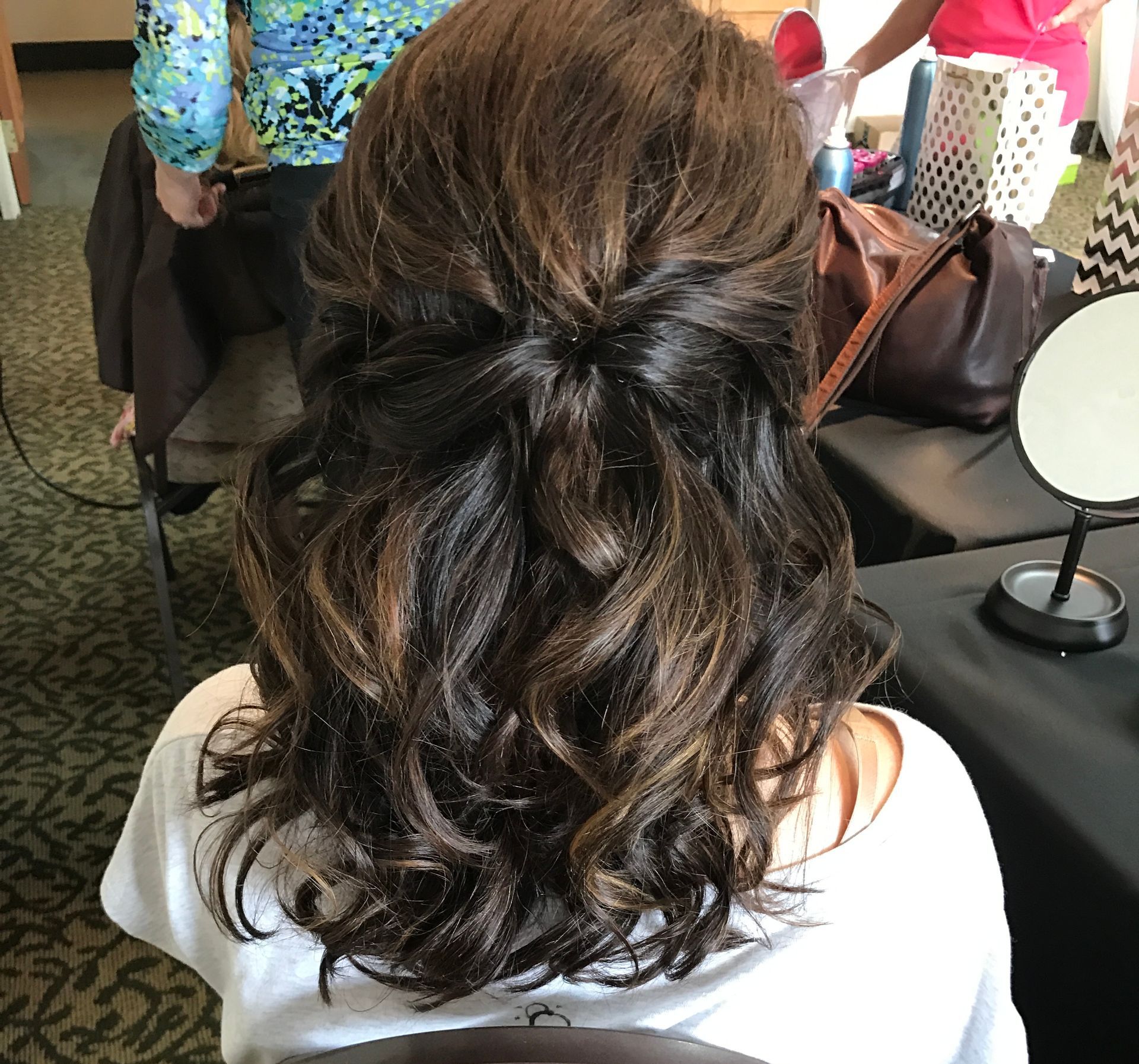 Woman with dark, wavy hair in a half-updo. Brown highlights, white shirt, sitting near a mirror.