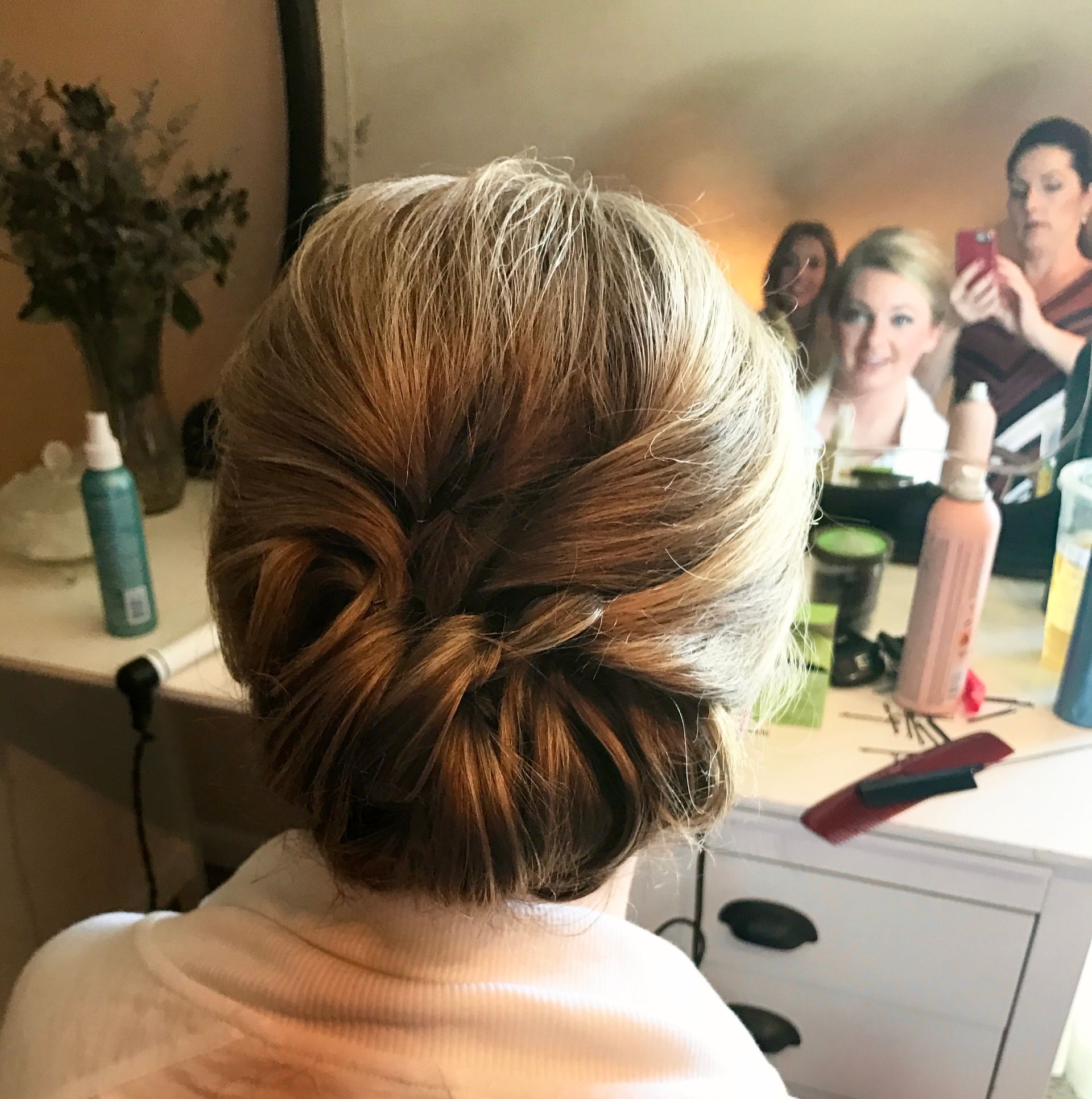 Woman's hair styled in a low bun. Friends in the mirror take photos. White vanity with sprays.