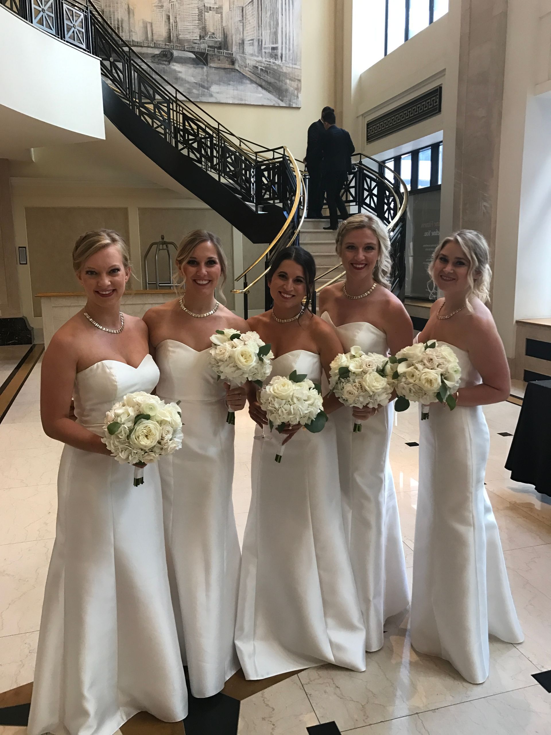 Bridesmaids in white strapless gowns holding bouquets, standing in front of a staircase.