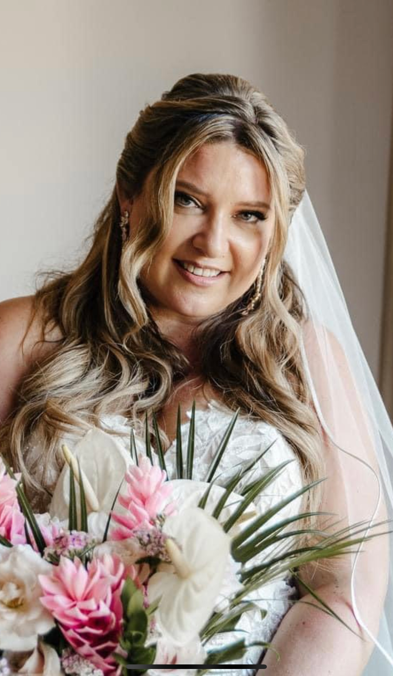 Bride smiling, holding a bouquet of pink and white flowers. Wearing a strapless wedding dress, veil, and updo.