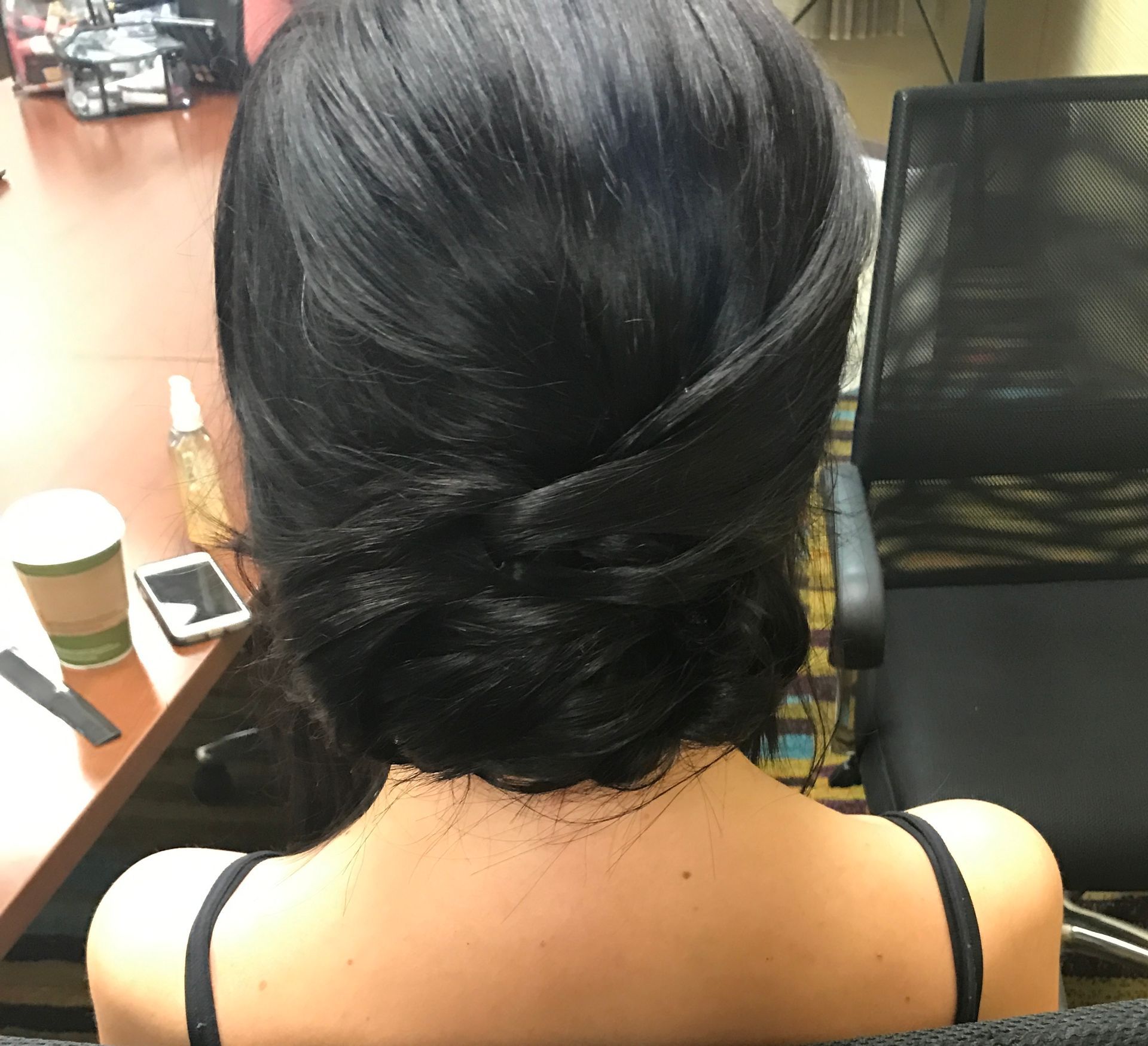 Woman with dark hair styled in a low bun, back view. She is wearing a black top, seated.