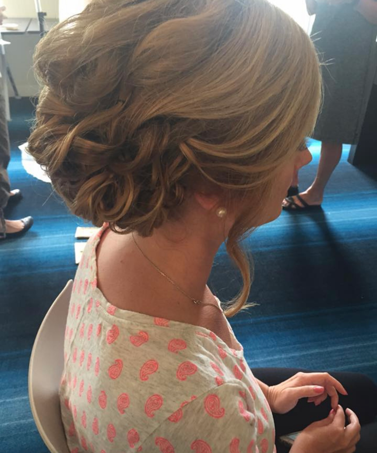 Woman with blonde updo hairstyle wearing a pink polka dot top, sitting indoors.