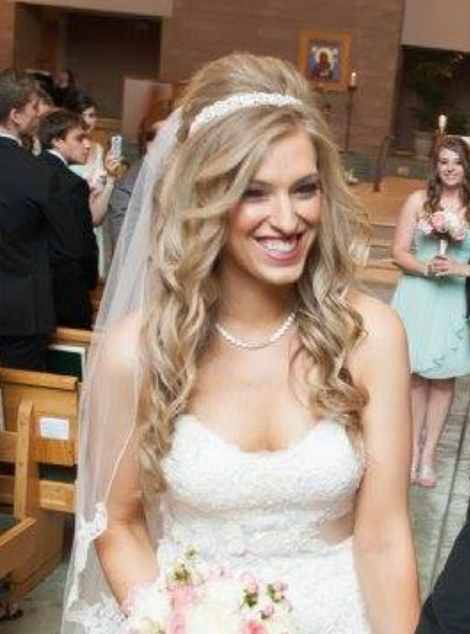 Bride in a white wedding dress, smiling, long wavy blonde hair, veil, pearl necklace, church setting.