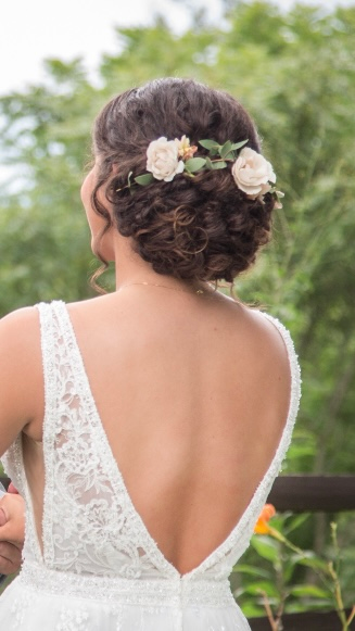 Bride with updo hairstyle, backless white lace dress, adorned with flowers. Outdoor setting.