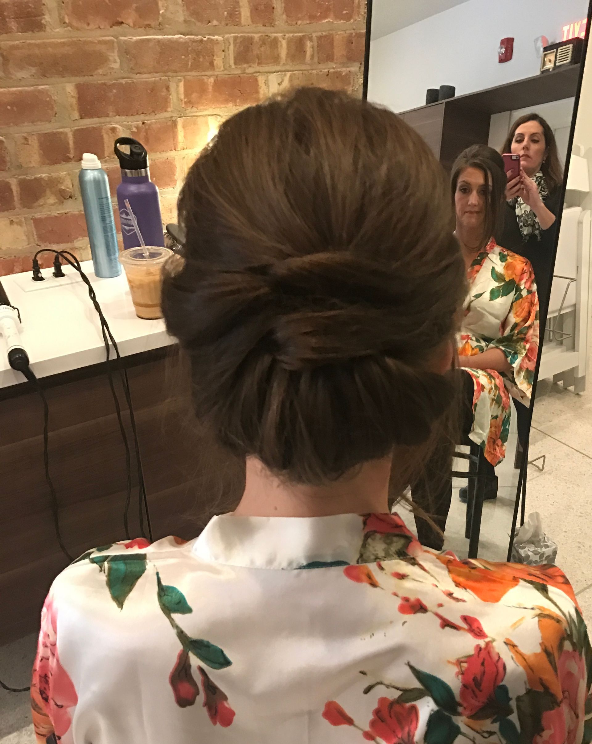 Woman with dark hair in a low bun, wearing a floral robe, views her reflection in a mirror.