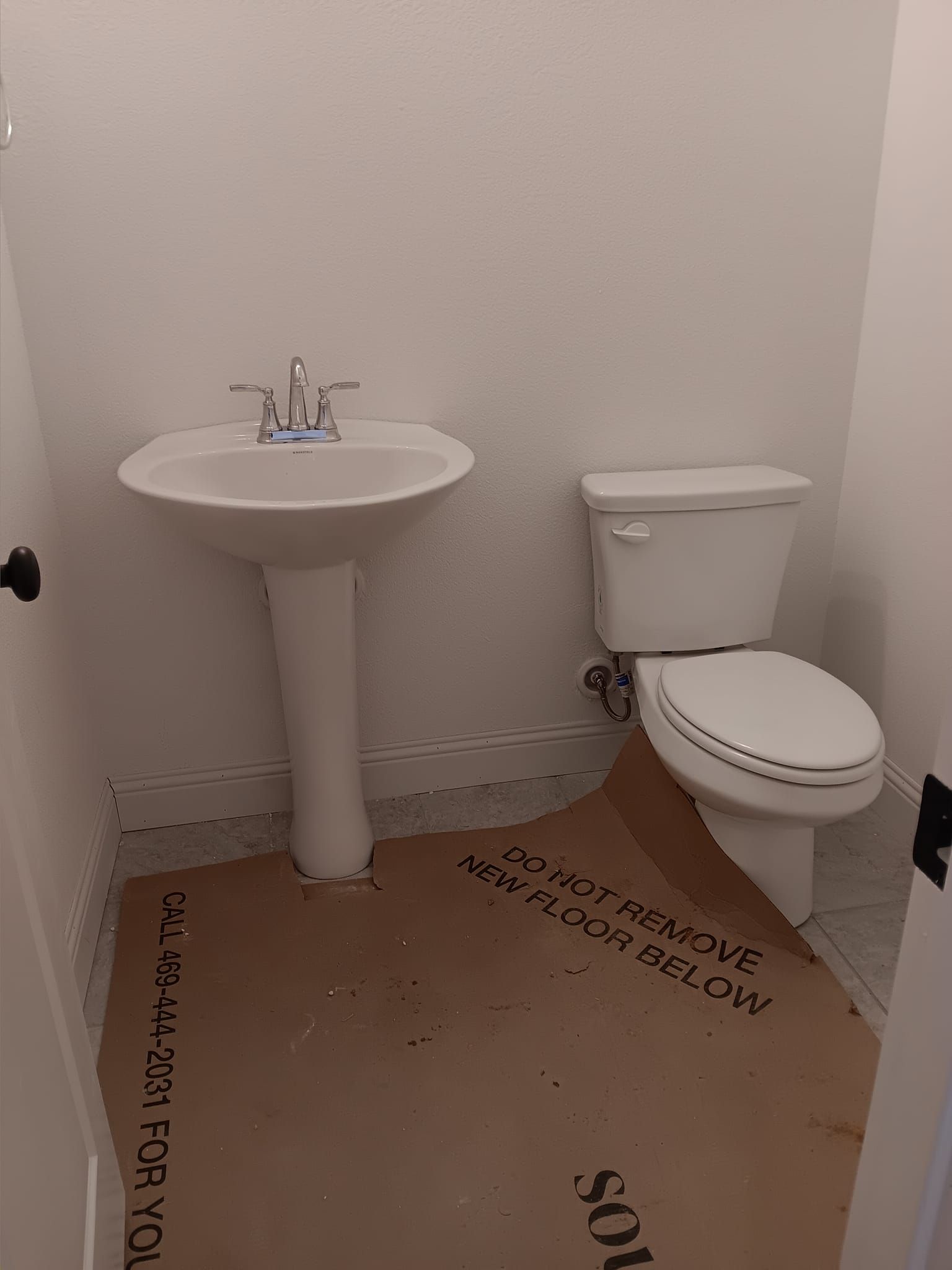 Small white bathroom with pedestal sink, toilet, and cardboard floor protection.