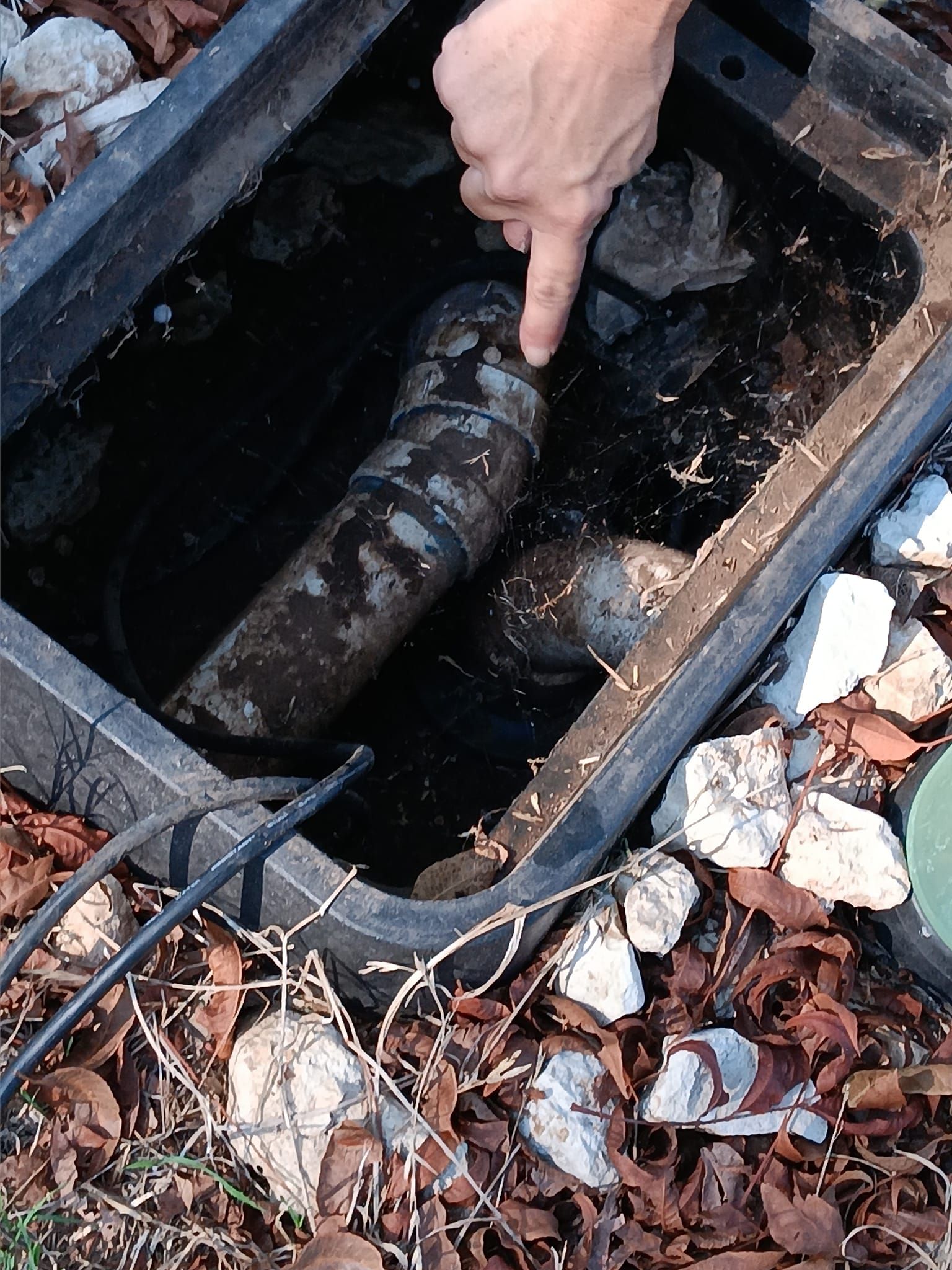 Hand pointing at an old bottle lying in a dark drainage hole with debris around it