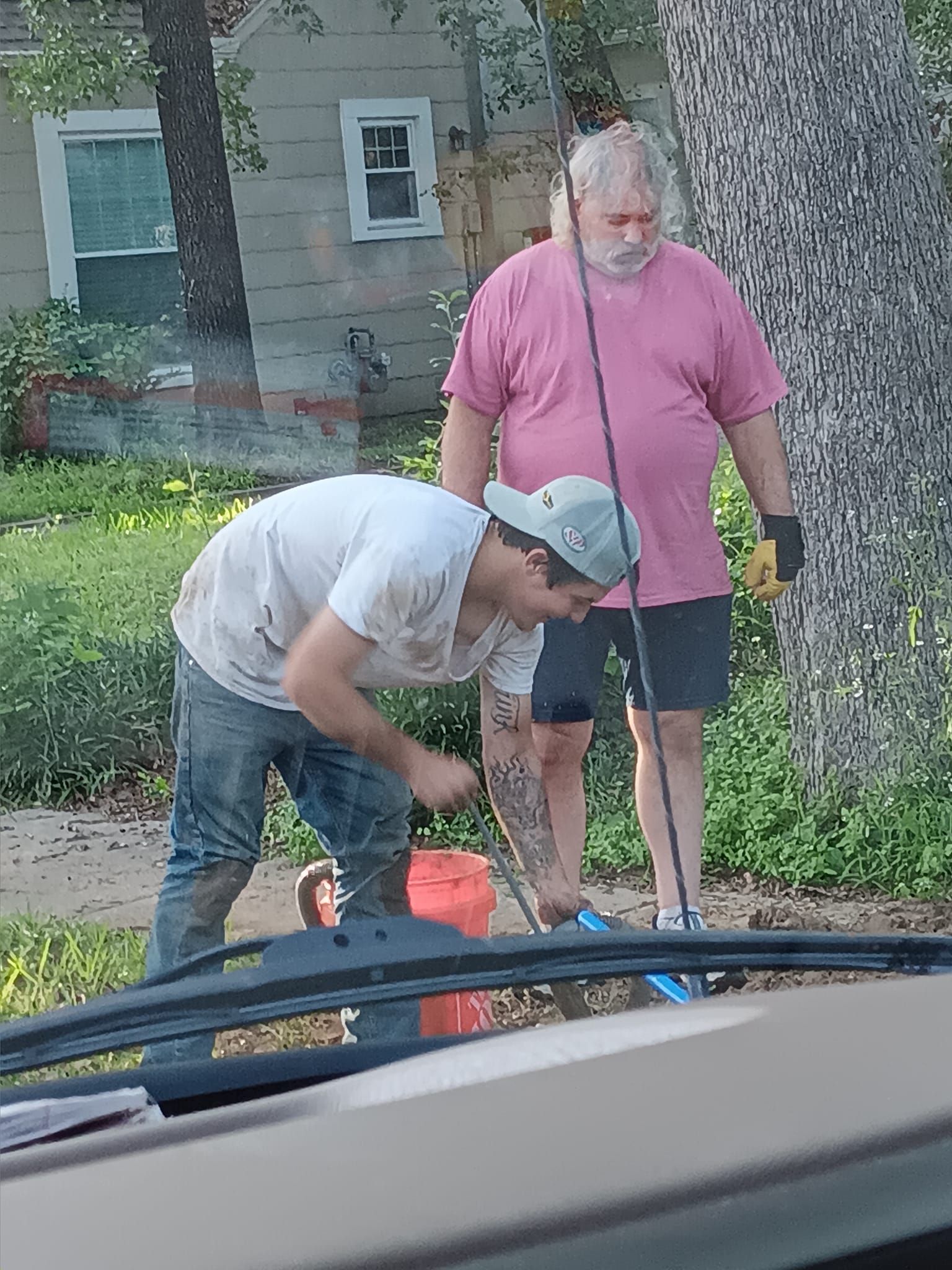 Two men work with equipment on a sidewalk near a house, one bending over an orange container.