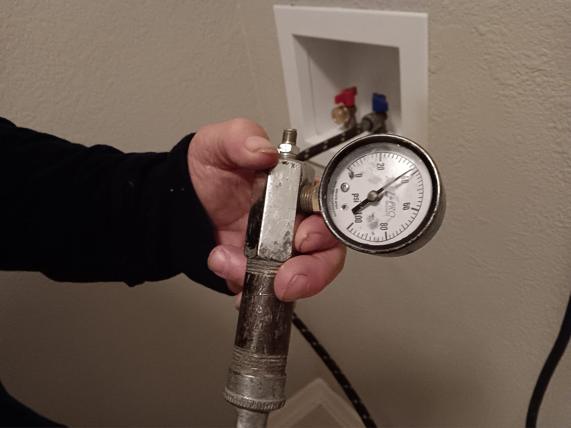 Hand holding a pressure gauge attached to a pipe near a wall valve outlet.