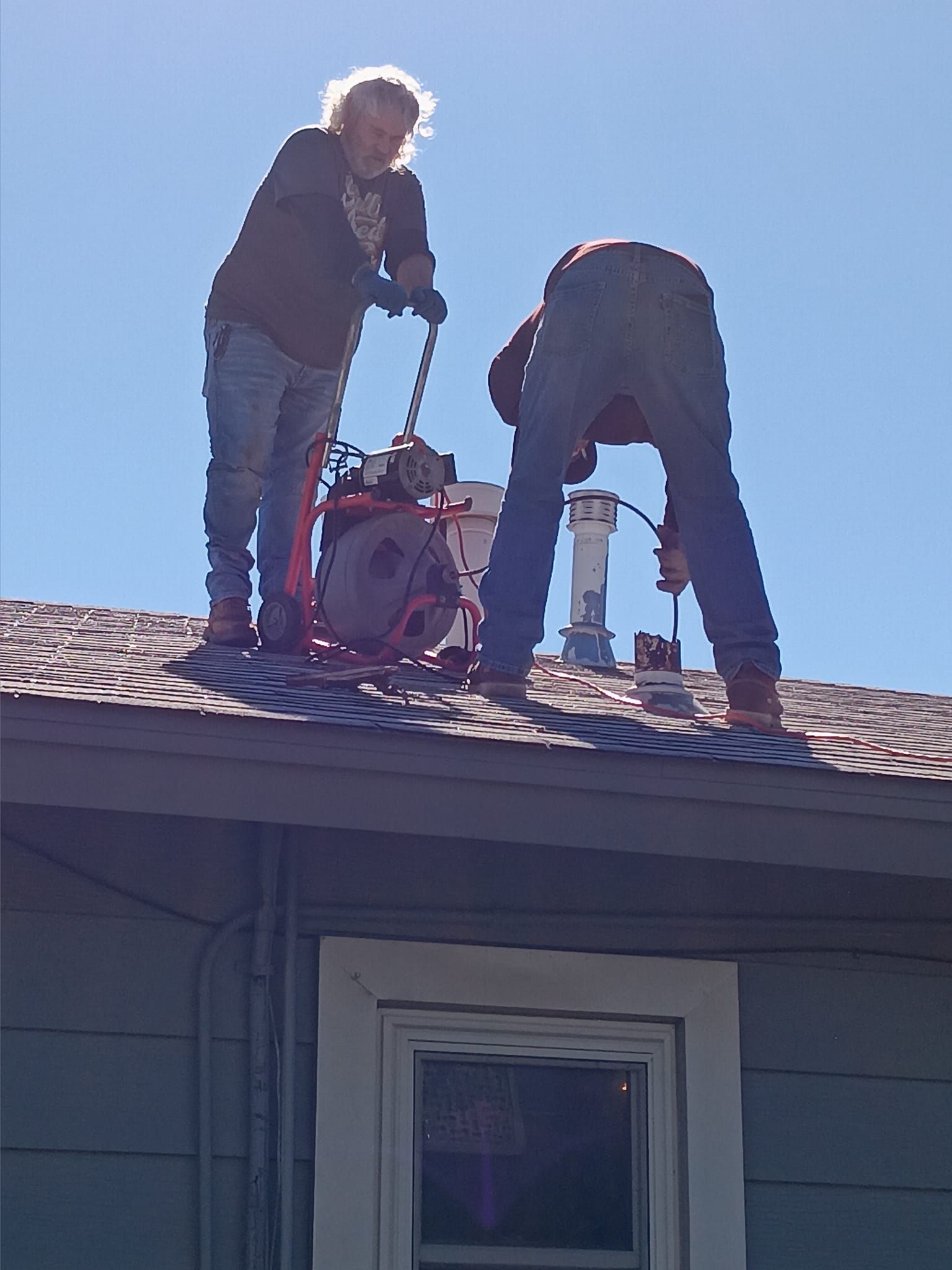 Two workers on a roof handling a red power tool in bright sunlight