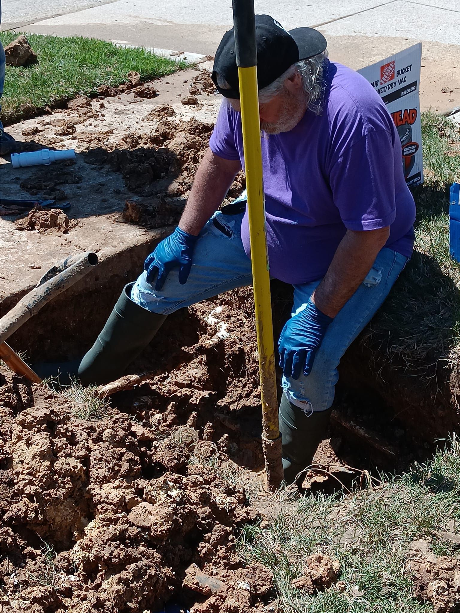 Worker in purple shirt and blue gloves digging dirt by a roadside trench with a shovel