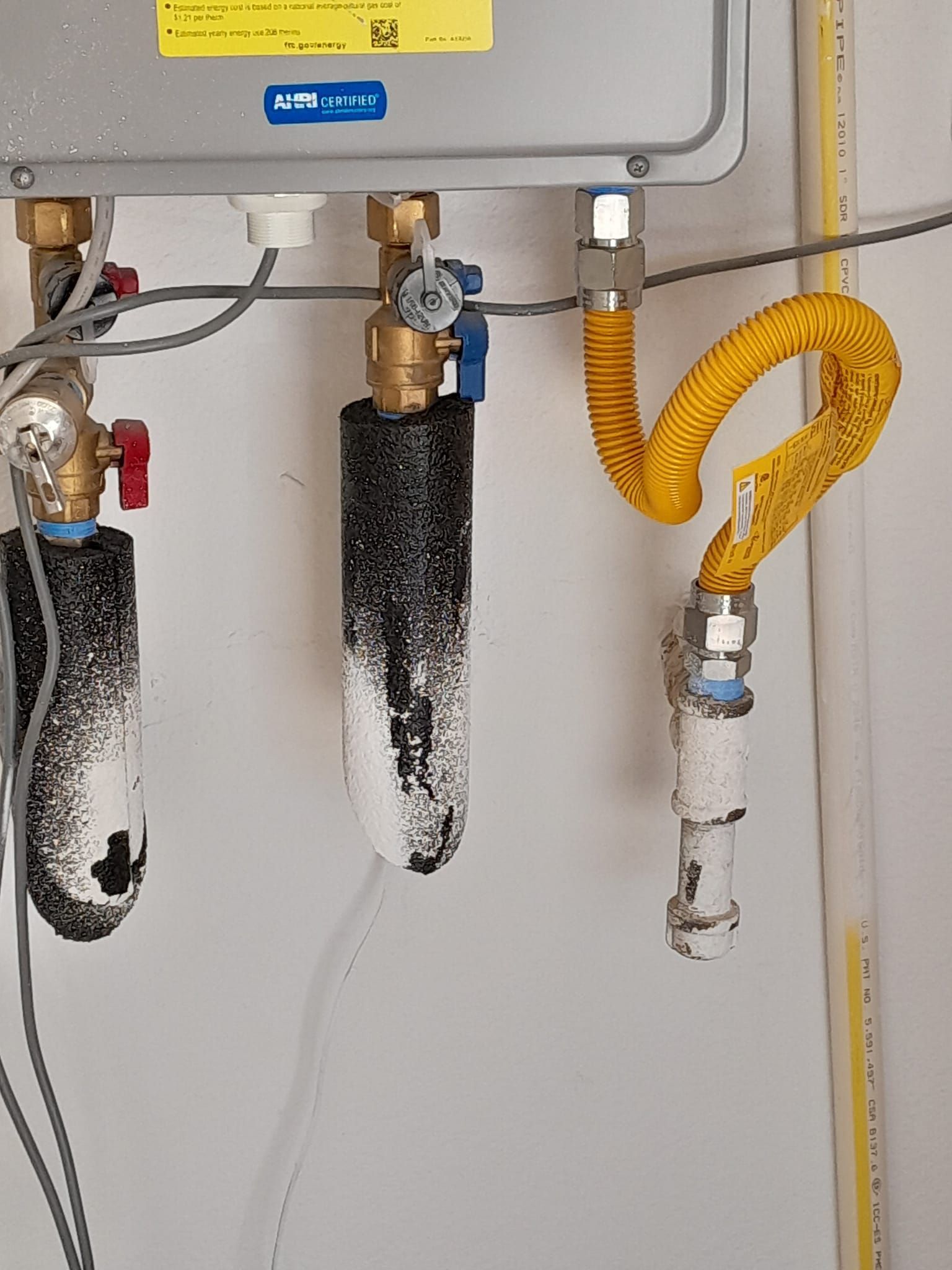 Water heater pipes and valves with insulation and a yellow flexible gas line in a utility room.