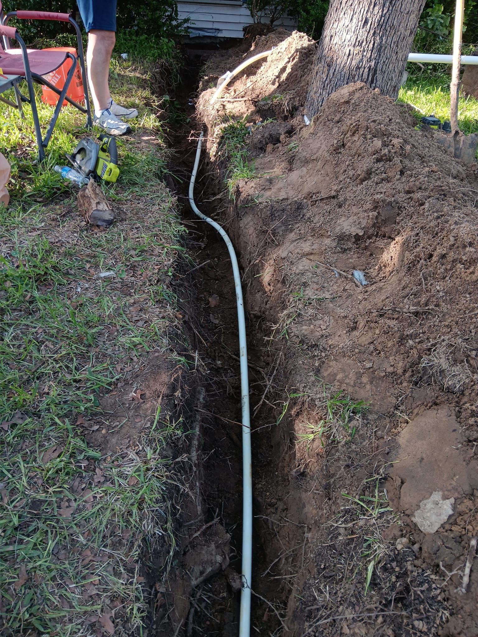 Person digging a narrow trench beside a large tree, with a white pipe running through the soil.