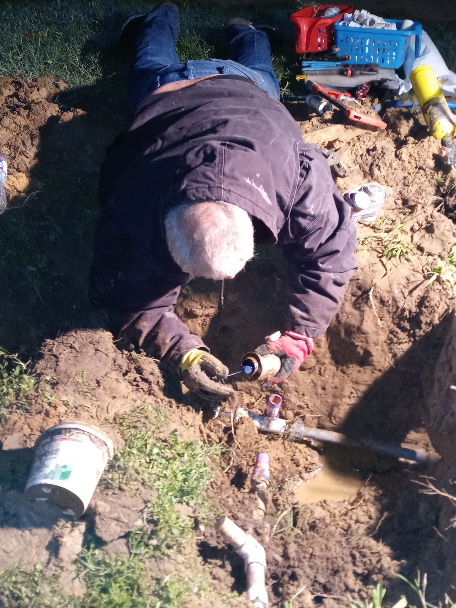 Person kneeling in a dirt trench, repairing a pipe with tools and a blue spray can nearby.