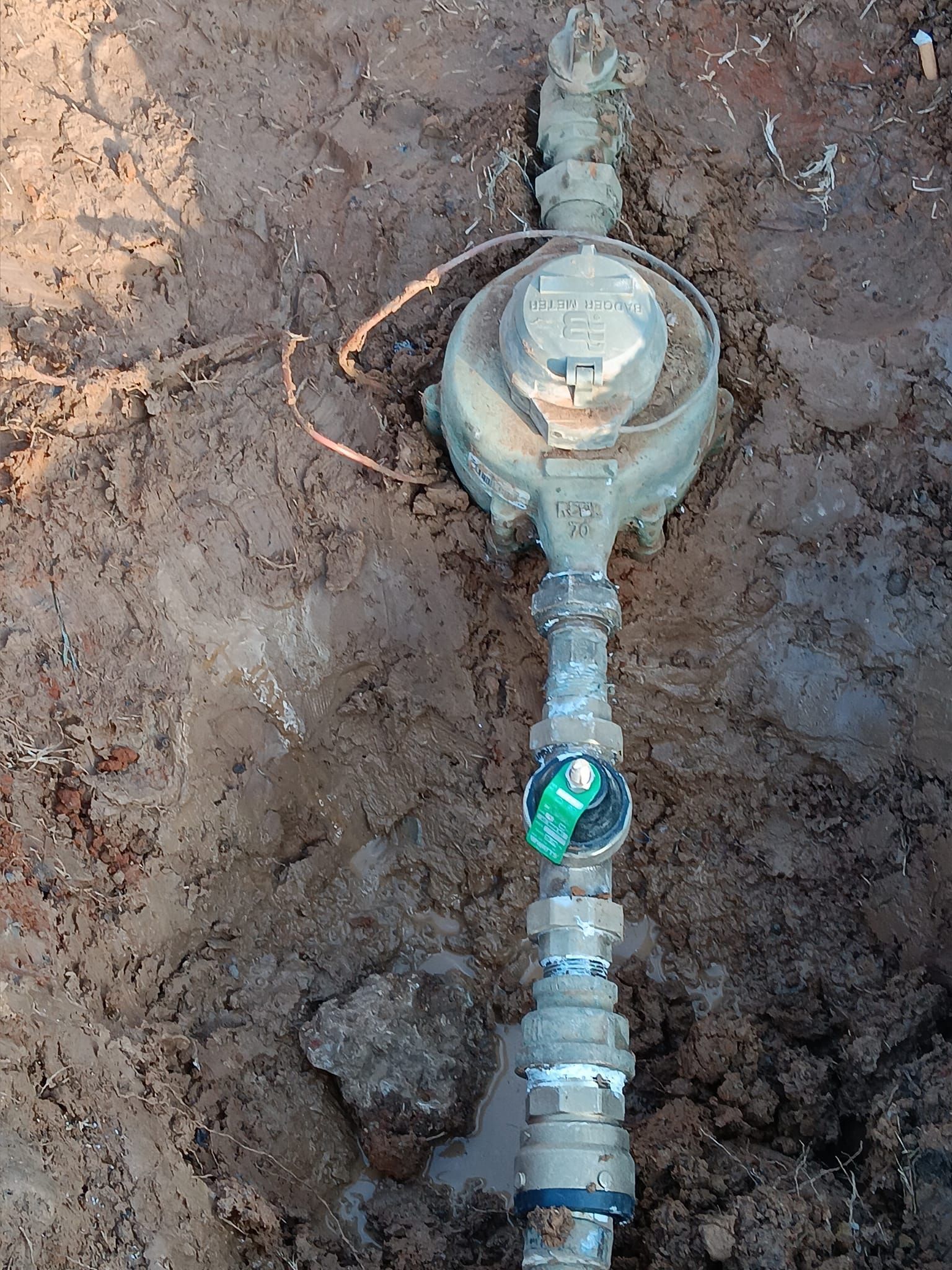 Water meter and underground pipe exposed in muddy dirt with a green valve.