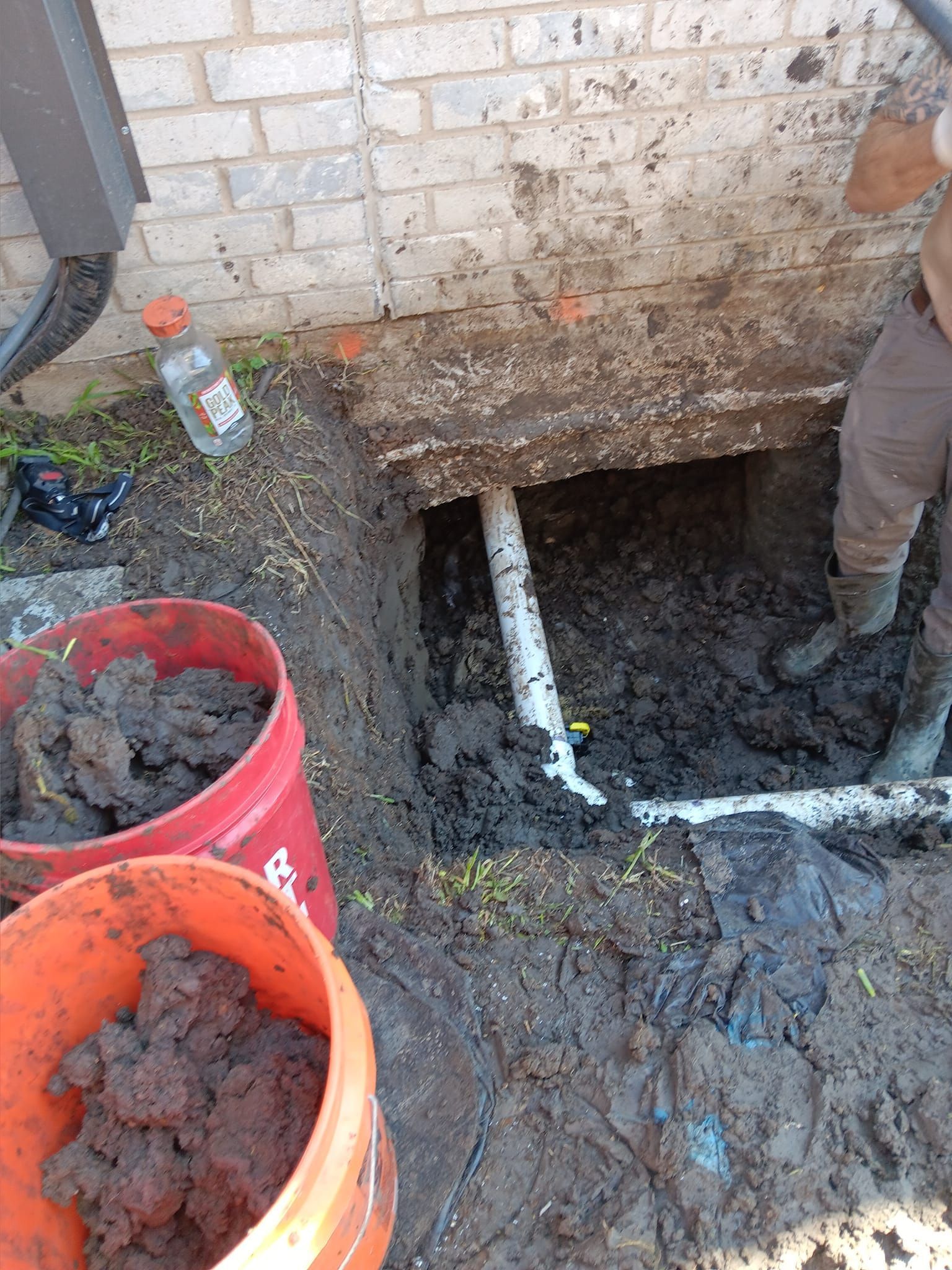 Mud-filled excavation beside a brick wall with exposed white pipe and orange buckets in the foreground