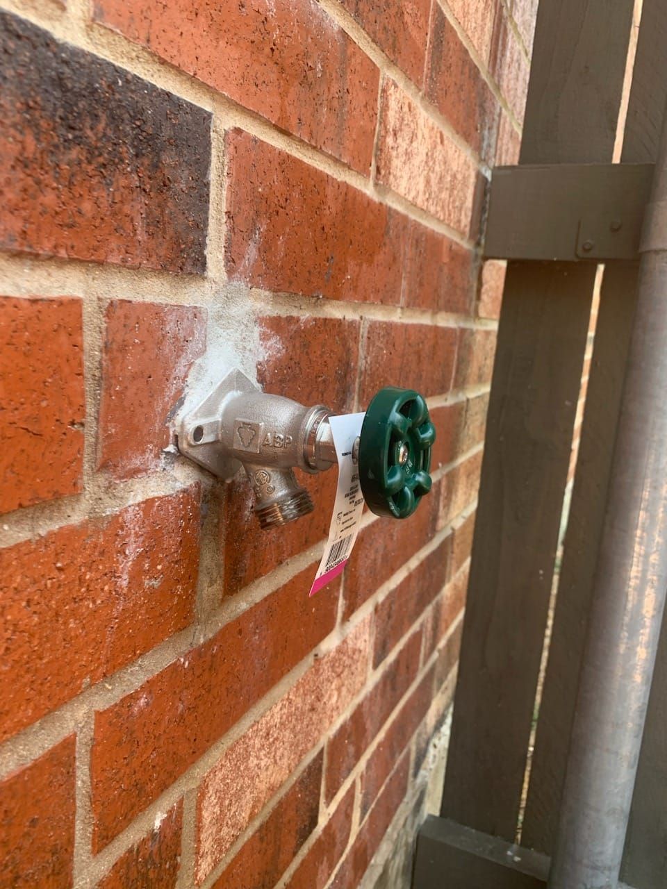 Green hose spigot protruding from a brick wall beside a wooden fence, with white sealant around the base