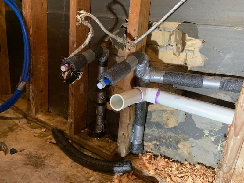 Exposed plumbing pipes and drains inside an unfinished wall with insulation and wood framing.