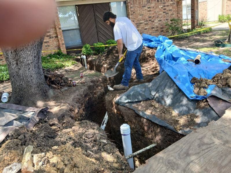 Worker digging near house foundation beside blue tarp and tree root, with exposed trench and debris