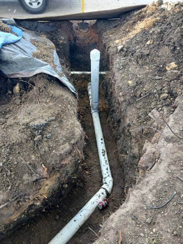 White PVC pipe in a dirt trench beneath a driveway edge, with an open vertical vent pipe.