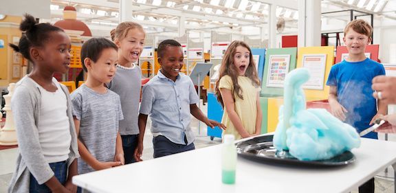A group of children are standing around a table looking at an elephant toothpaste experiment.