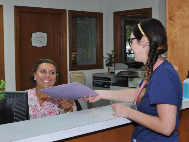 Two women at a desk exchanging documents. One smiles, wearing a floral shirt, the other, in blue scrubs, is handing over a file.