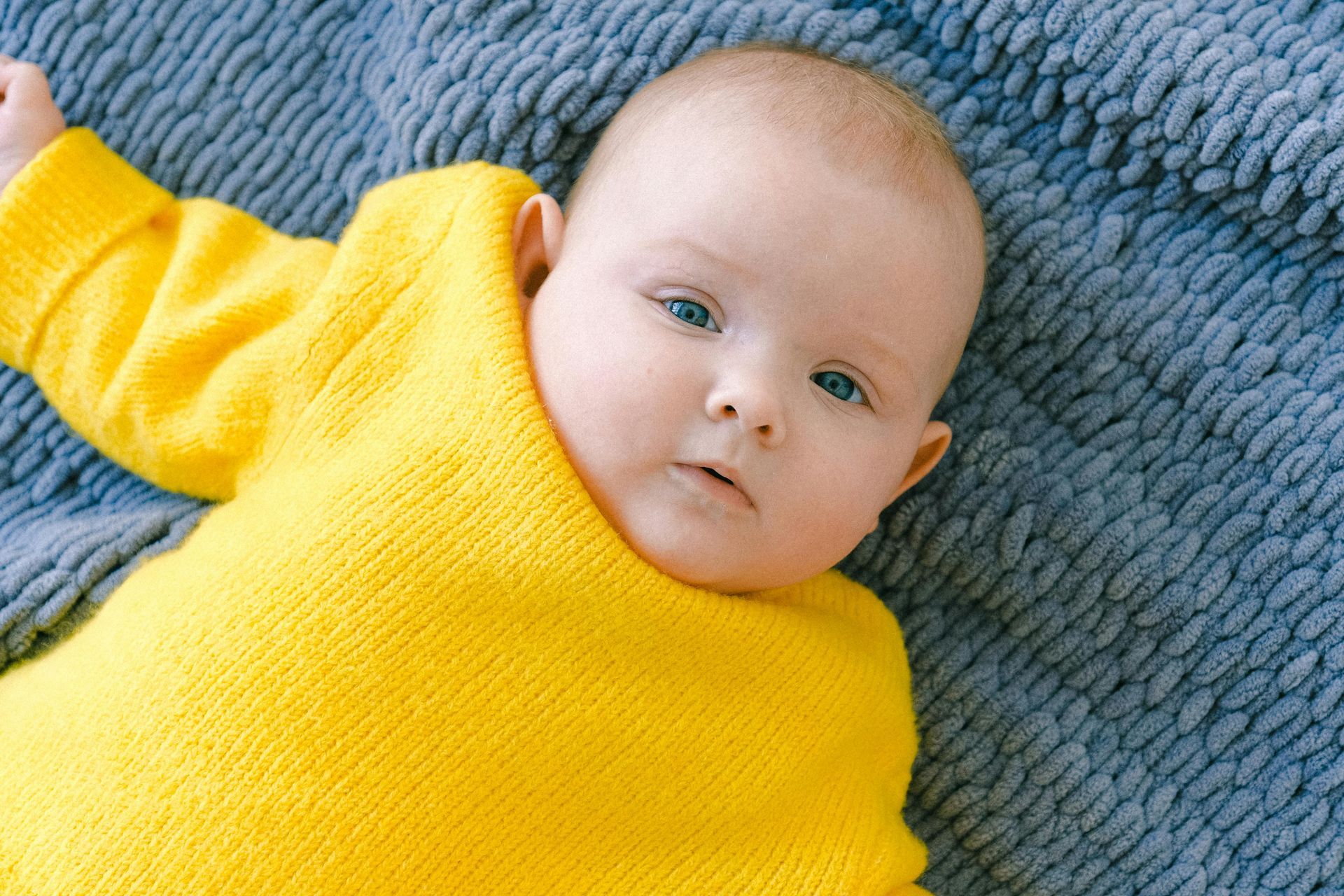 A baby in a bright yellow sweater is laying on the ground looking at the camera.