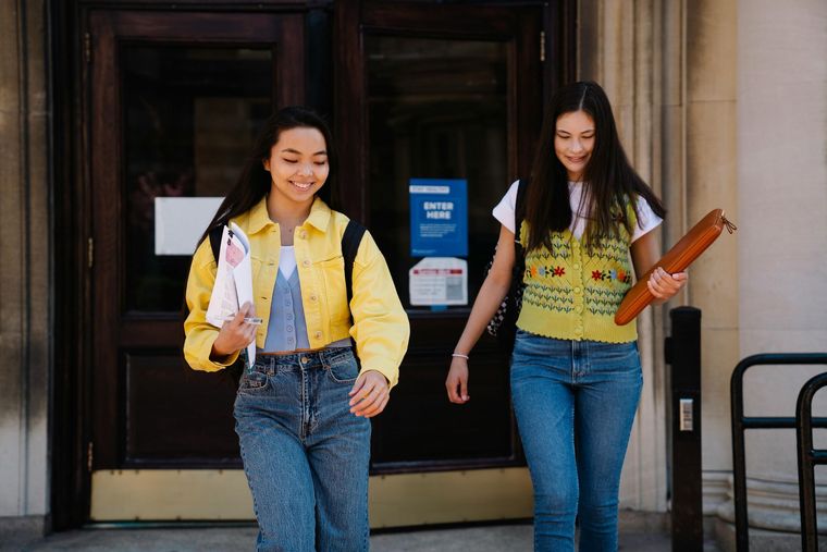 Two smiling young women exiting a building, one with a yellow jacket and the other with a yellow sweater vest.