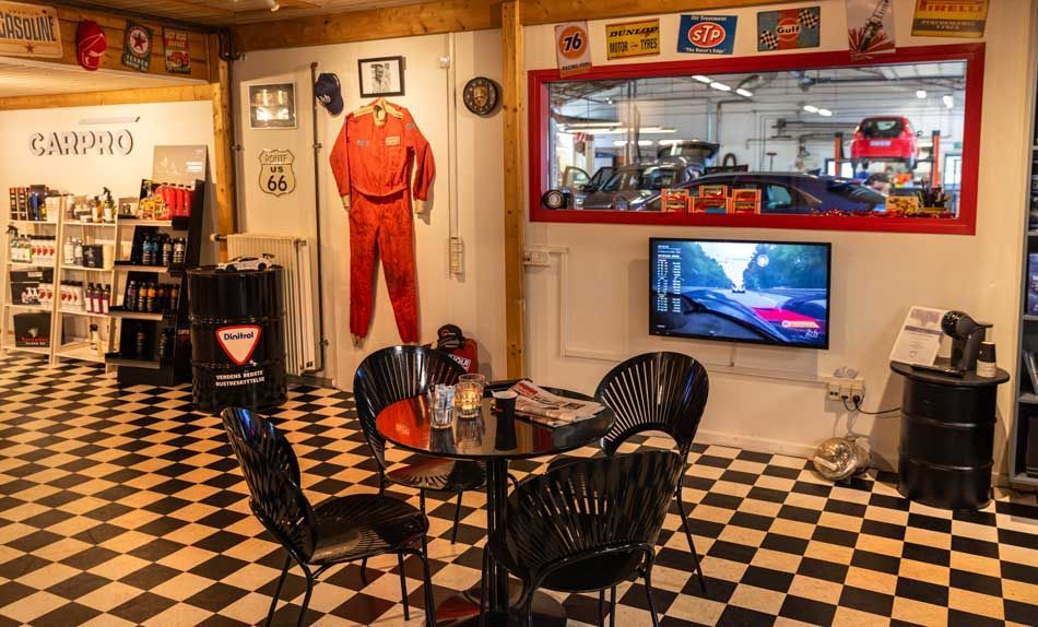 Et venteområde med biltema, sort bord, stole og et skakbrætgulv. Et vindue har udsigt over et bilværksted.