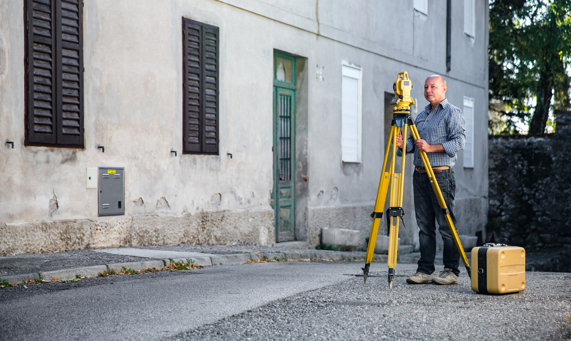 A person stands on a paved surface next to a building, operating a yellow surveying total station on a tripod.