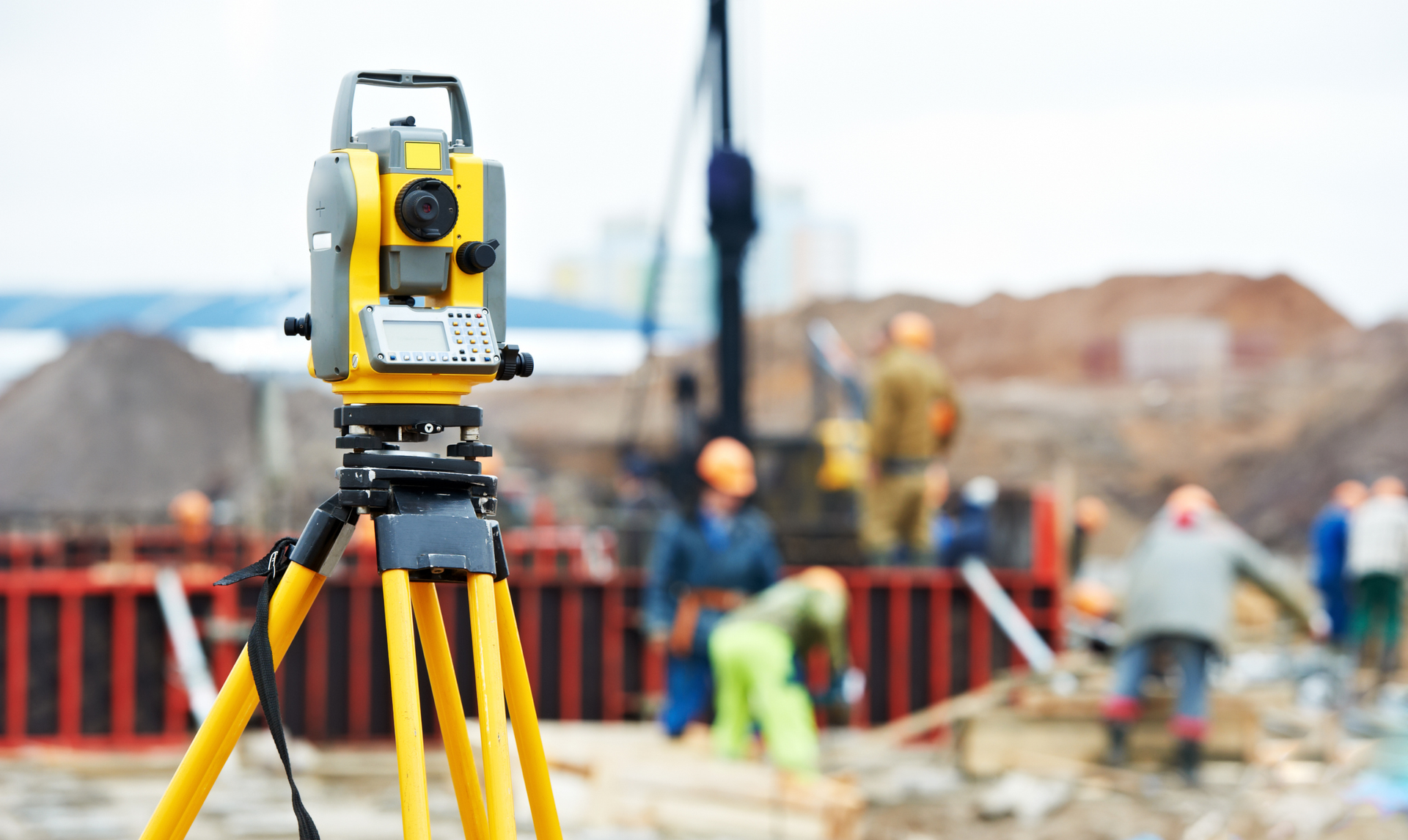 A yellow total station on a tripod in the foreground, with construction workers and activity in a blurred background.