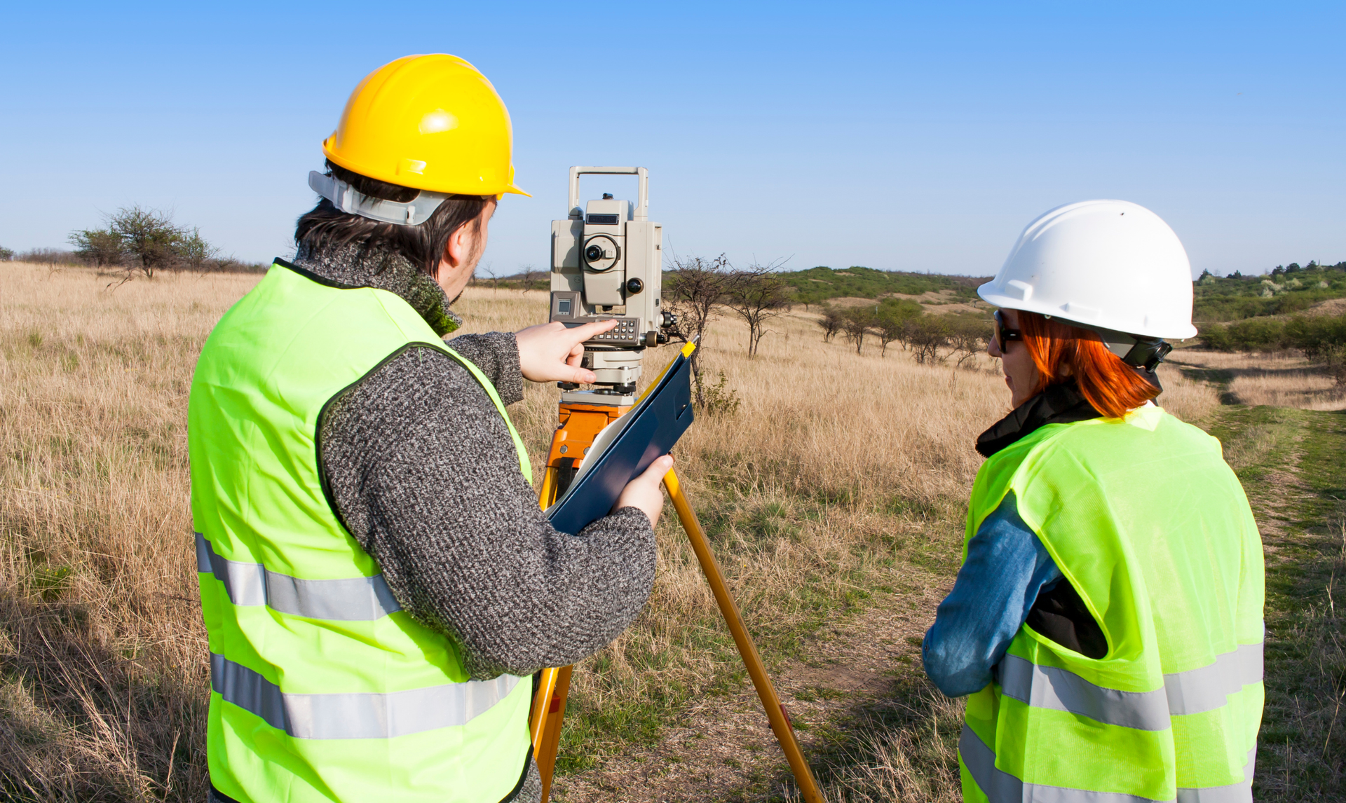 Two land surveyors in high-visibility vests and hard hats work with a theodolite in a dry, grassy outdoor field.
